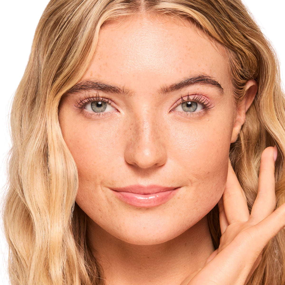 Model wearing ColourPop Fresh Kiss Glossy Balm in Strawberry Milk, showcasing a sheer pink glossy lip finish; full-face studio close-up with natural makeup, soft waves, and a hand at her cheek against a white background.