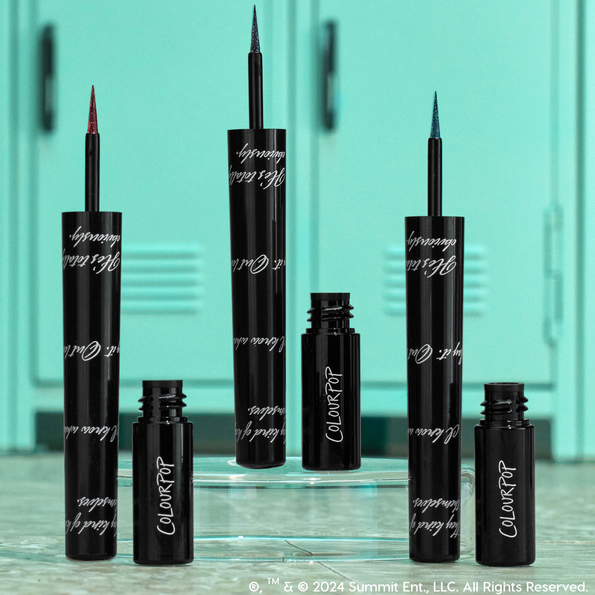 ColourPop Graphix Ink Liner I Know What You Are stands uncapped, showing ultra-fine red, blue, and teal shimmer tips; glossy black tubes with white script sit on a clear riser before mint school lockers for a back-to-school vibe.