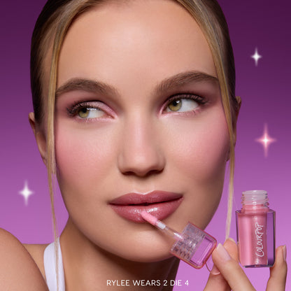Model applies Gloss Me Up ultra glossy lip set in Heart to Get, holding the pink shimmery ColourPop-style tube against a purple starry background, showcasing high-shine nude-pink lips and matching soft glam makeup for a glossy finish.