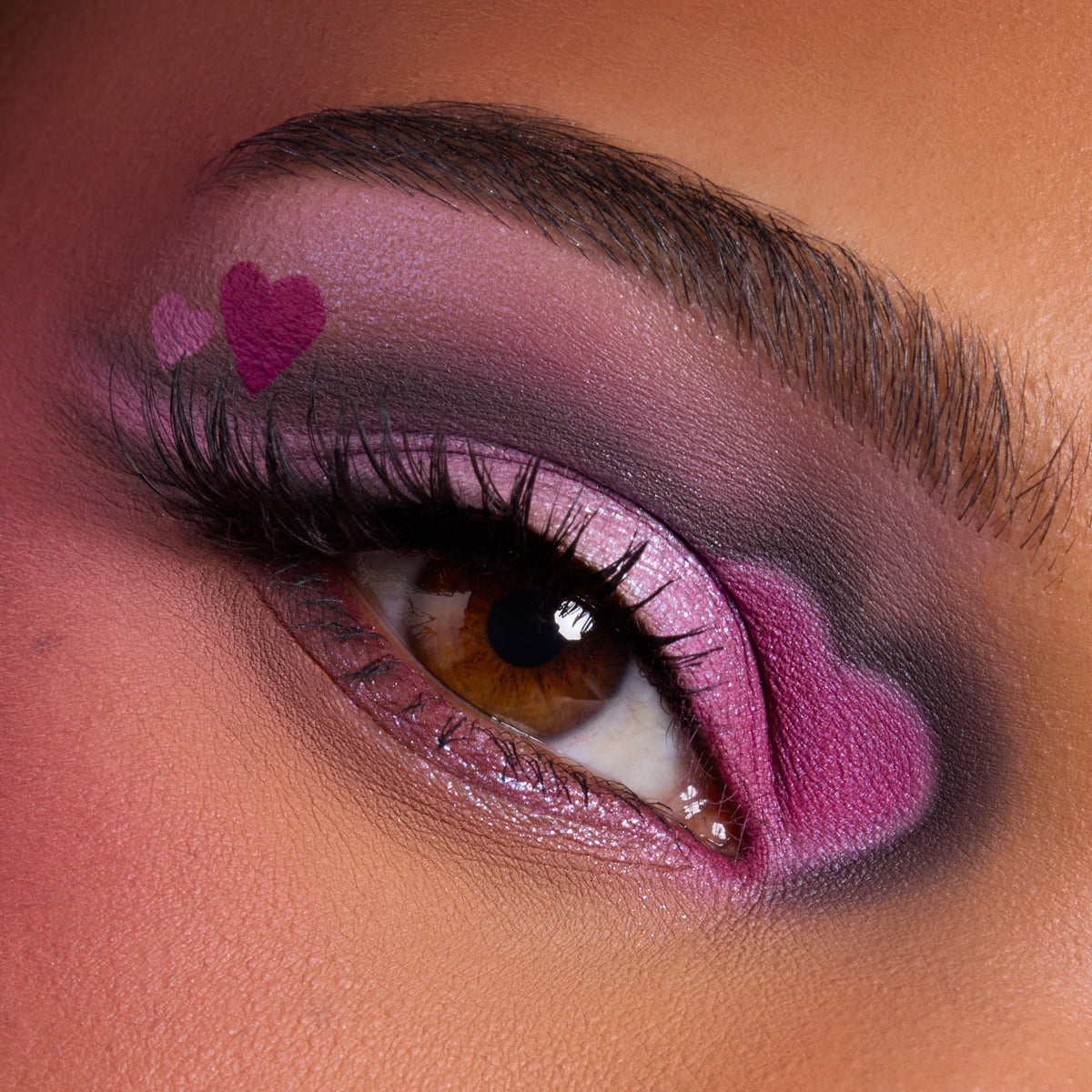 Close-up of a brown eye wearing the Heart to Get shadow palette, featuring bold pink and purple shimmer with heart-shaped accents at the inner and outer corners, showcasing dramatic, romantic eye makeup on warm-toned skin.