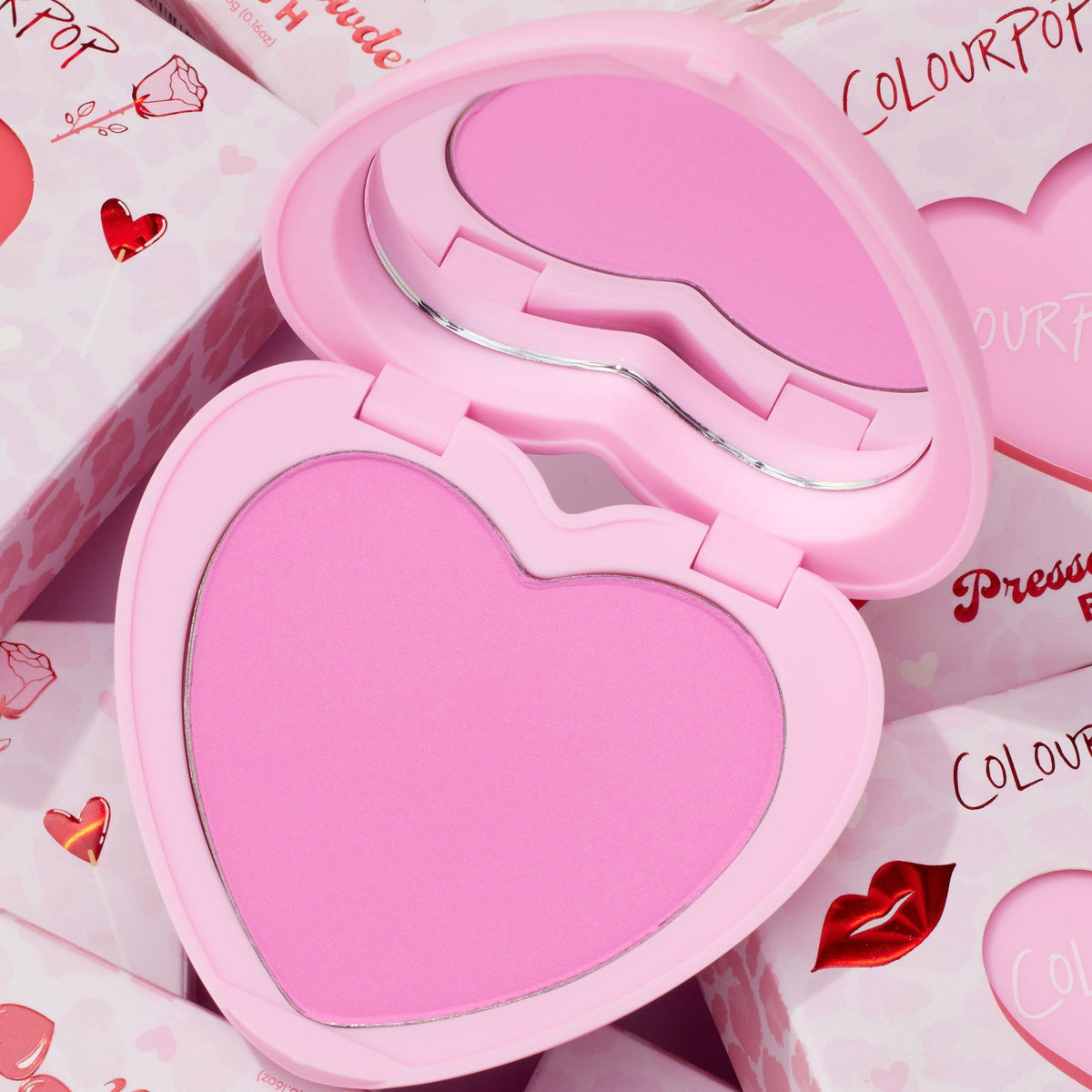 Heart-shaped Hot 2 Touch compact opens to reveal bright pink ColourPop Pressed Powder Blush, set in a pastel pink case with built-in mirror, resting atop Valentine-themed boxes with hearts and red lip graphics.