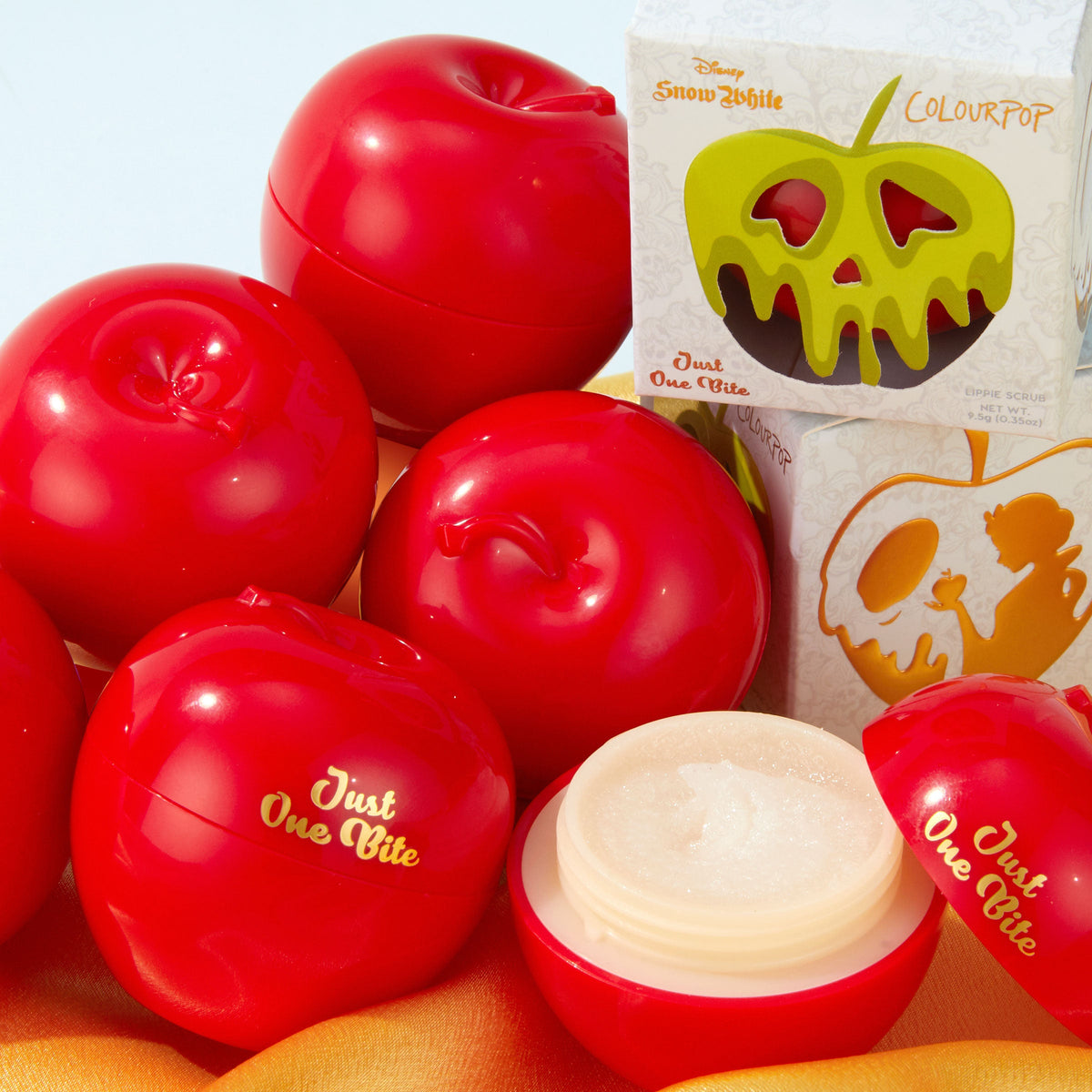 Just One Bite ColourPop Lip Scrub opens to reveal a sugary exfoliant in a glossy red apple-shaped jar, surrounded by matching apple containers and Disney Snow White packaging with a green poison-apple graphic; 0.3 oz lip scrub.