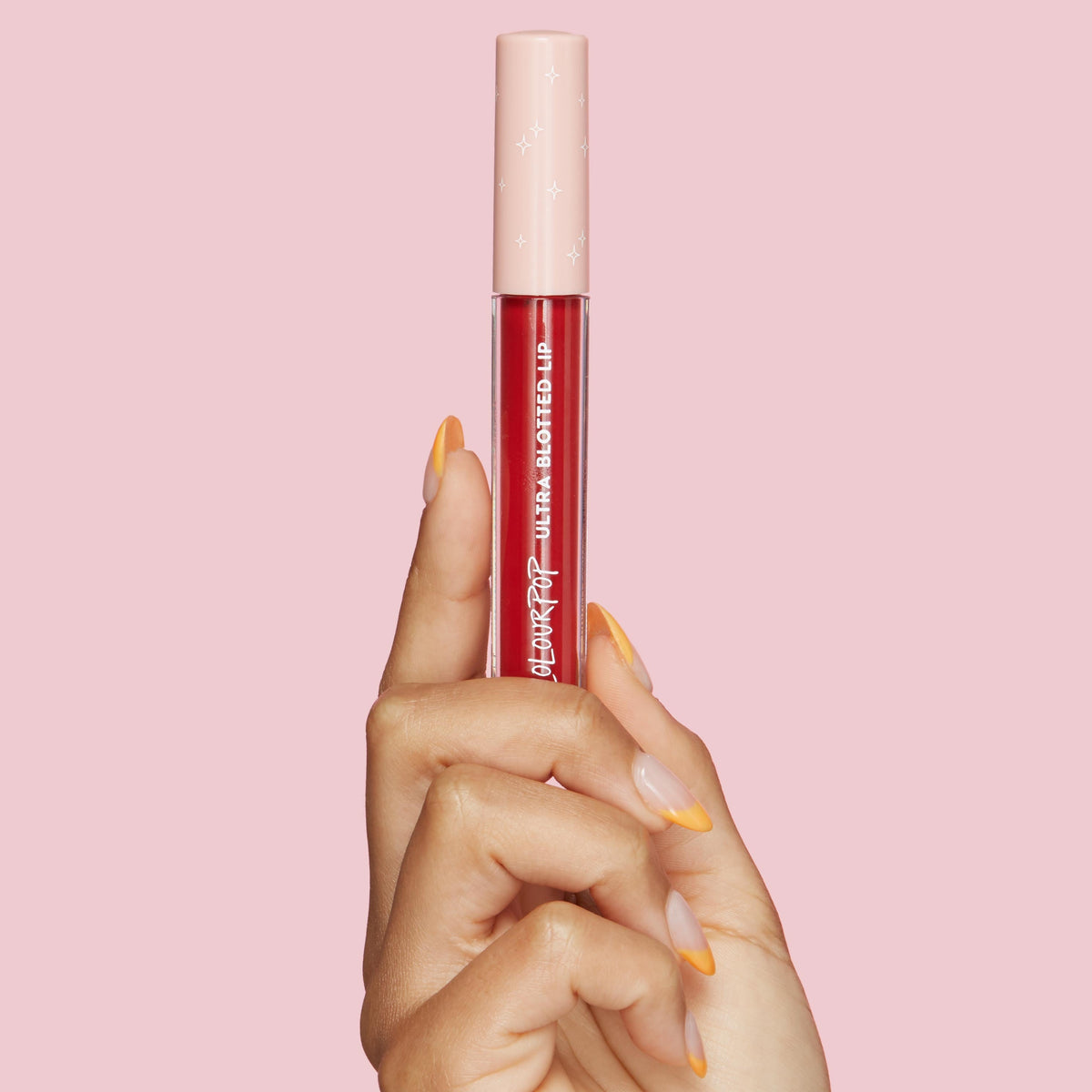 ColourPop Ultra Blotted Lip in Keep It Classy held upright by a hand with orange French tips, showcasing a clear tube of deep berry-red tint with a blush pink cap dotted with stars against a solid pink background.
