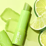 Key Lime ColourPop Lip Balm stick uncapped and twisted up, resting on a glossy swatch with the cap beside it, styled top-down on a lime-green background with fresh key lime wedges emphasizing the bright citrus flavor.