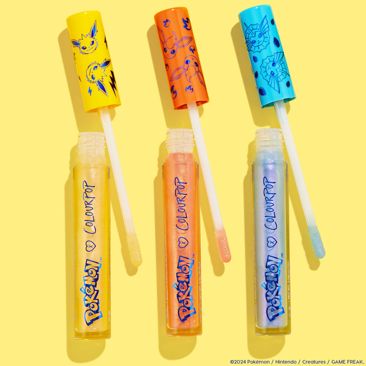 ColourPop Ultra Glossy Lip Evolution - three shimmery lip glosses with doe‑foot wands out - displayed in a flat lay on a bright yellow backdrop, in Pokémon x ColourPop packaging with Jolteon, Flareon, and Vaporeon art; yellow, orange, and blue hues.