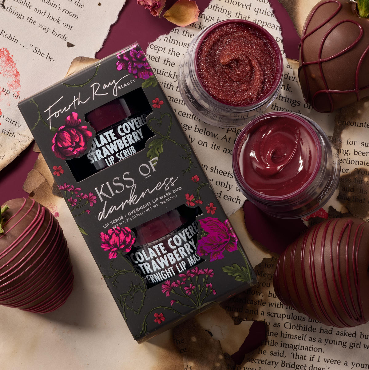 Fourth Ray Beauty Kiss of Darkness lip care kit shows chocolate-covered-strawberry lip scrub and overnight lip mask, jars open to reveal shimmery berry textures, beside the floral black box, styled with chocolate-dipped strawberries on torn book pages; ColourPop Lip Care Kit.