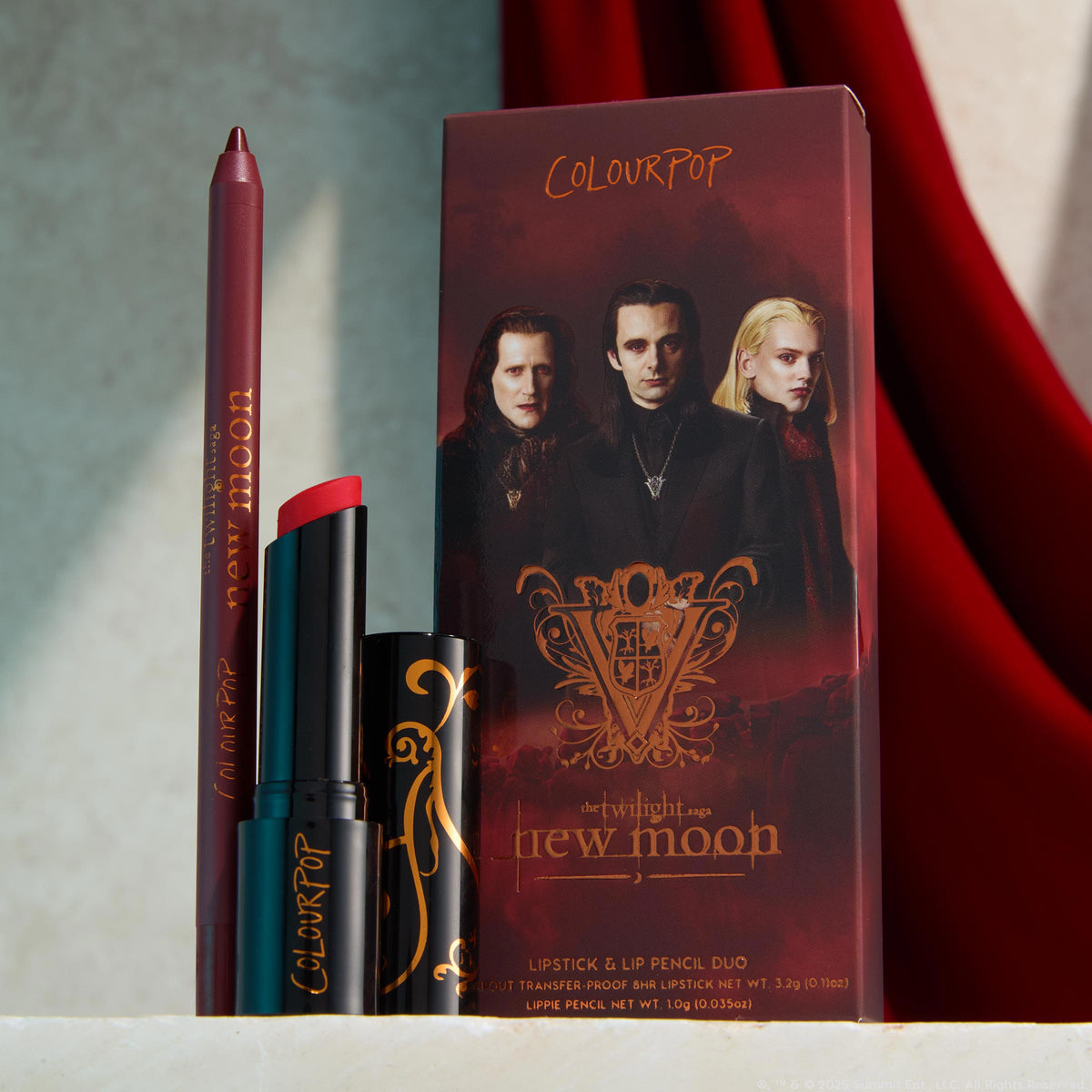ColourPop You Can't Trust Vampires Power Pout Lipstick stands uncapped to reveal a deep red bullet beside a plum lip pencil, set before a Twilight New Moon Volturi-themed box on a light stone surface with a red velvet backdrop.