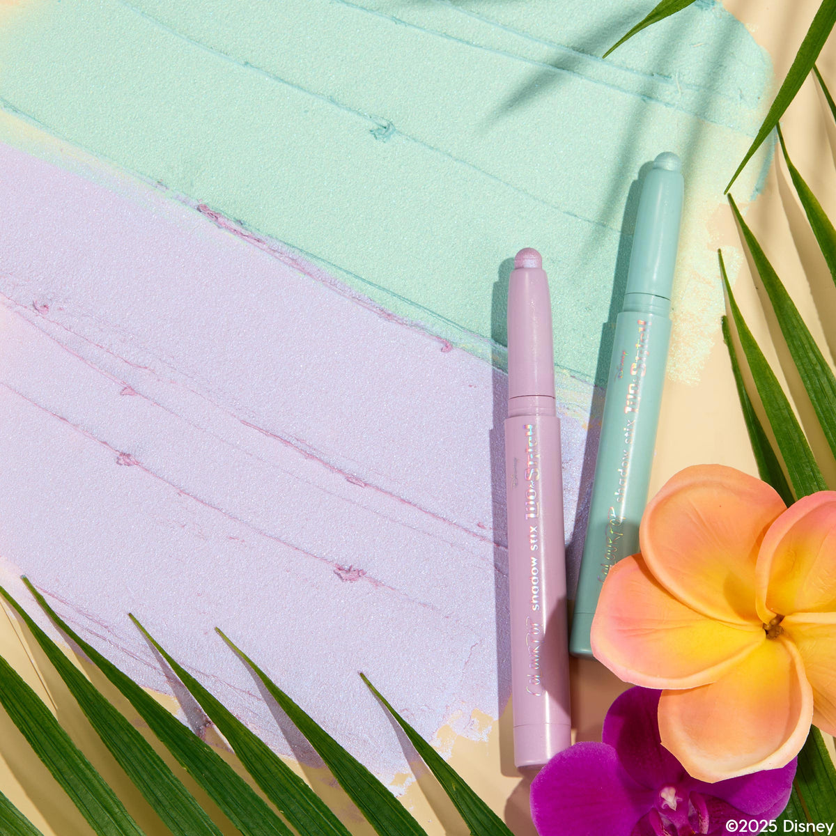 Two pastel ColourPop Shadow Stix, Cute and Feisty, in mint and lilac, rest on matching shimmery swatches, framed by tropical palm leaves and pink-orange plumeria and purple orchid, showcasing the creamy twist-up eyeshadow duo.