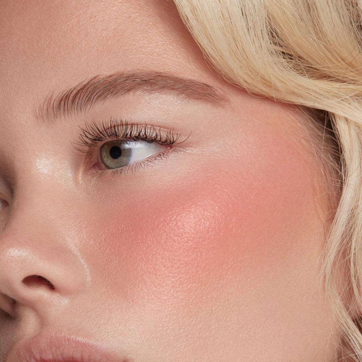 ColourPop Liquid Blush in Super Shy sweeping a dewy peach‑pink flush across the cheekbone, blended to a soft luminous sheen; extreme close-up shows natural lashes and brow with blonde strands framing the face.