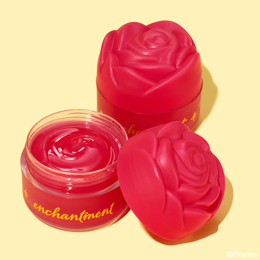 Enchanted Rose ColourPop Lip Mask shown open, revealing a swirled glossy raspberry-pink balm, flanked by two sculpted rose lids; red frosted glass jar with gold “enchantment” text on a pale yellow background.
