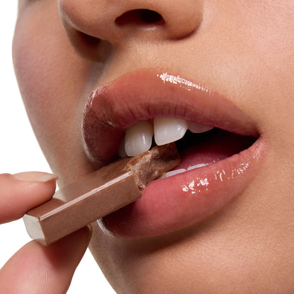 Glossy, hydrated lips bite a milk chocolate bar in a close-up beauty shot, highlighting the Triple Chocolate ColourPop Lip Care Kit and its lip mask shine.