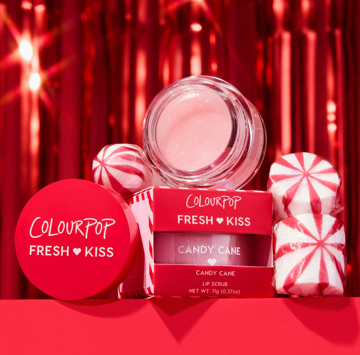 ColourPop Fresh Kiss Candy Cane lip scrub jar and red box showcasing a shimmery pink formula, staged with peppermint candies against a festive red tinsel backdrop; holiday lip exfoliator, 11 g (0.37 oz).