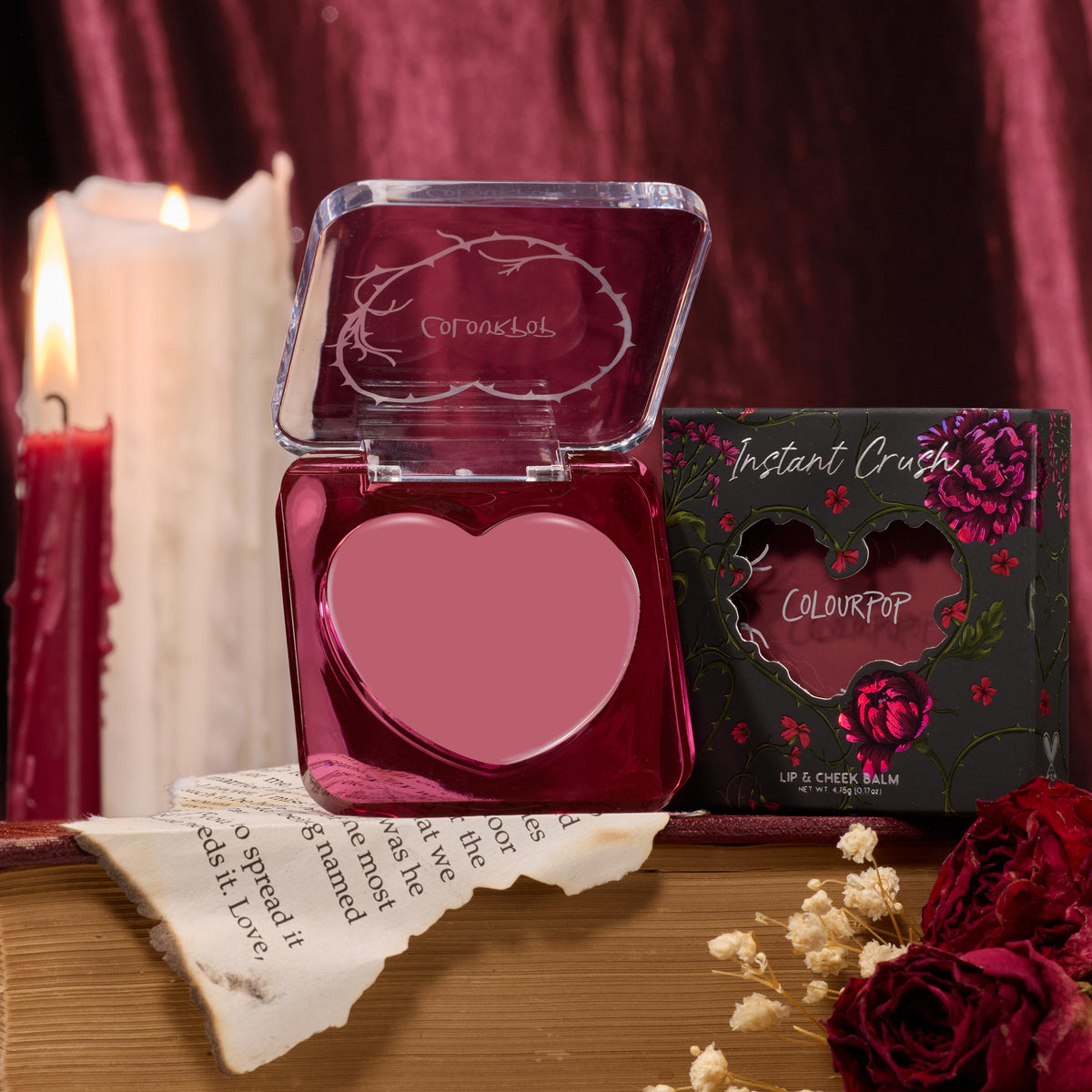 Heart-shaped Love Interest compact open to reveal rosy mauve balm, displayed with floral “Instant Crush” box, lit candles, a torn book page, and dried roses on a wood table - ColourPop Lip and Cheek Balm for lips and cheeks.
