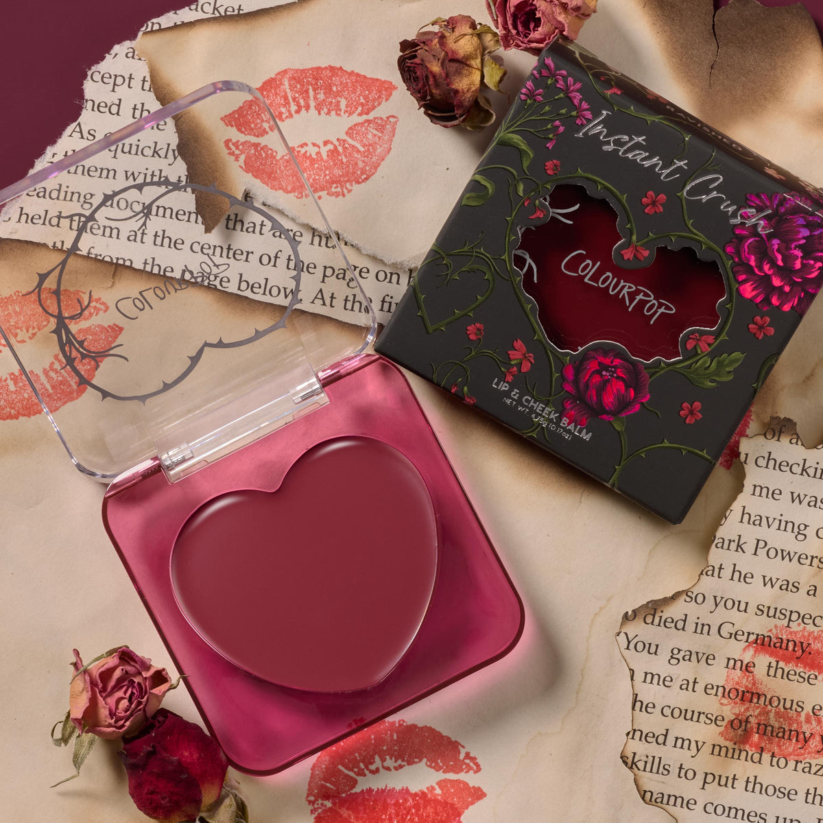 Ravished ColourPop Lip and Cheek Balm sits open, revealing a heart-shaped mauve tint in a translucent pink compact, beside its floral black box with heart window, styled on torn pages with lipstick kisses and dried roses.