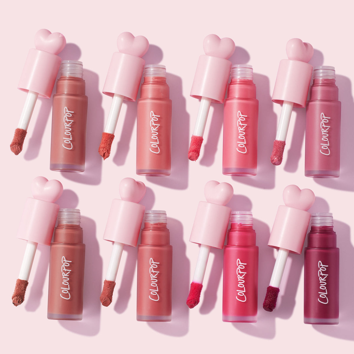 IKTR ColourPop Liquid Blush wands fanned out, doe-foot applicators coated in rosy, coral, and berry hues, with frosted tubes and pink heart caps arranged in a neat flat lay on a pale pink background, highlighting shade range and creamy texture.