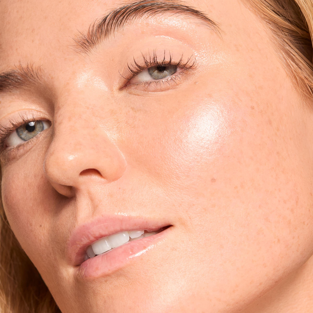 Cheekbone wearing ColourPop Liquid Highlighter in Sugar Donut catching light with a dewy pearlescent glow, shown in a tight macro of a model’s face with natural skin and glossy lips to showcase its reflective finish.