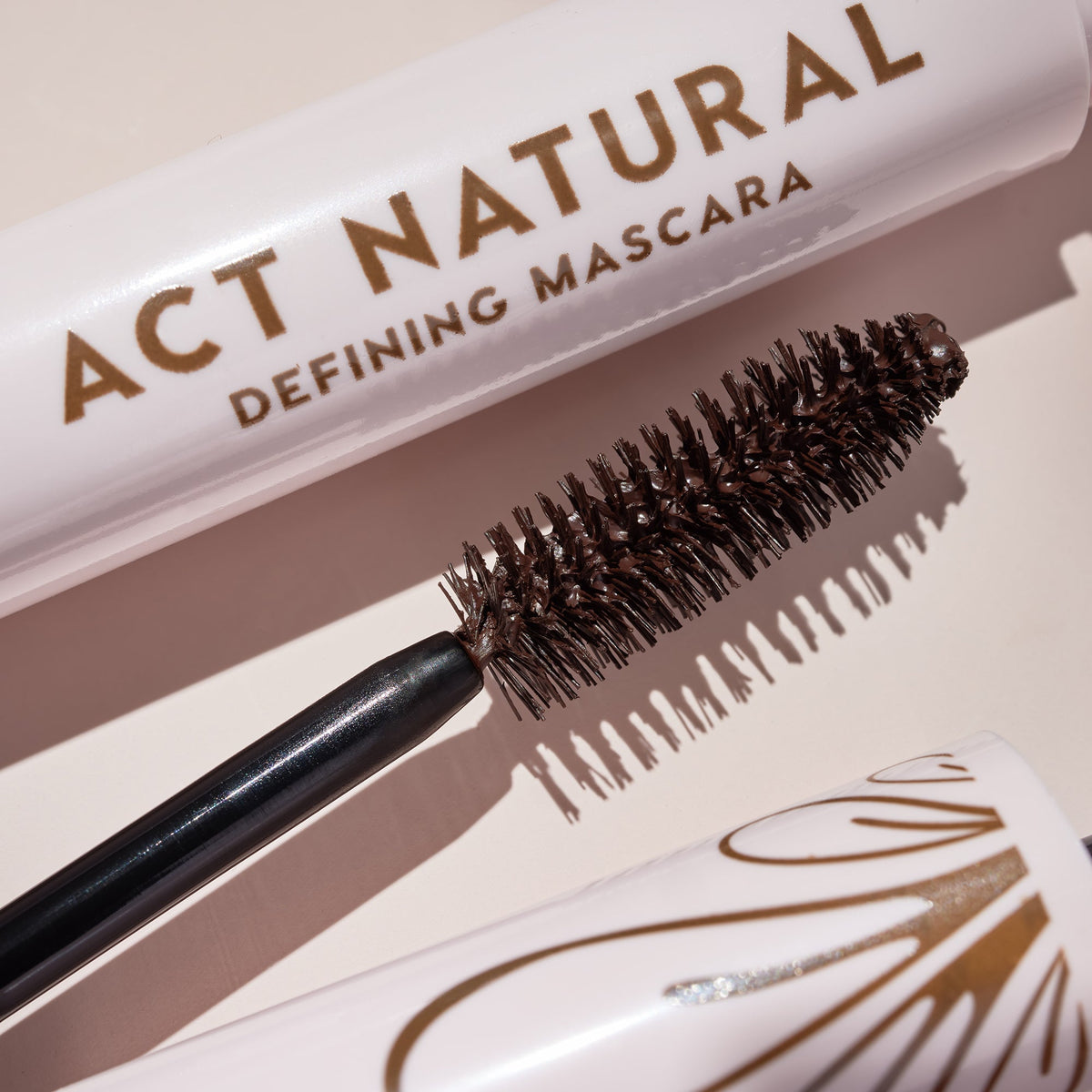 Mascara wand coated in rich brown formula rests beside a soft pink tube reading Act Natural Defining Mascara, highlighting dense spoolie bristles; ColourPop Act Natural Mascara in Brown shown on a neutral backdrop.