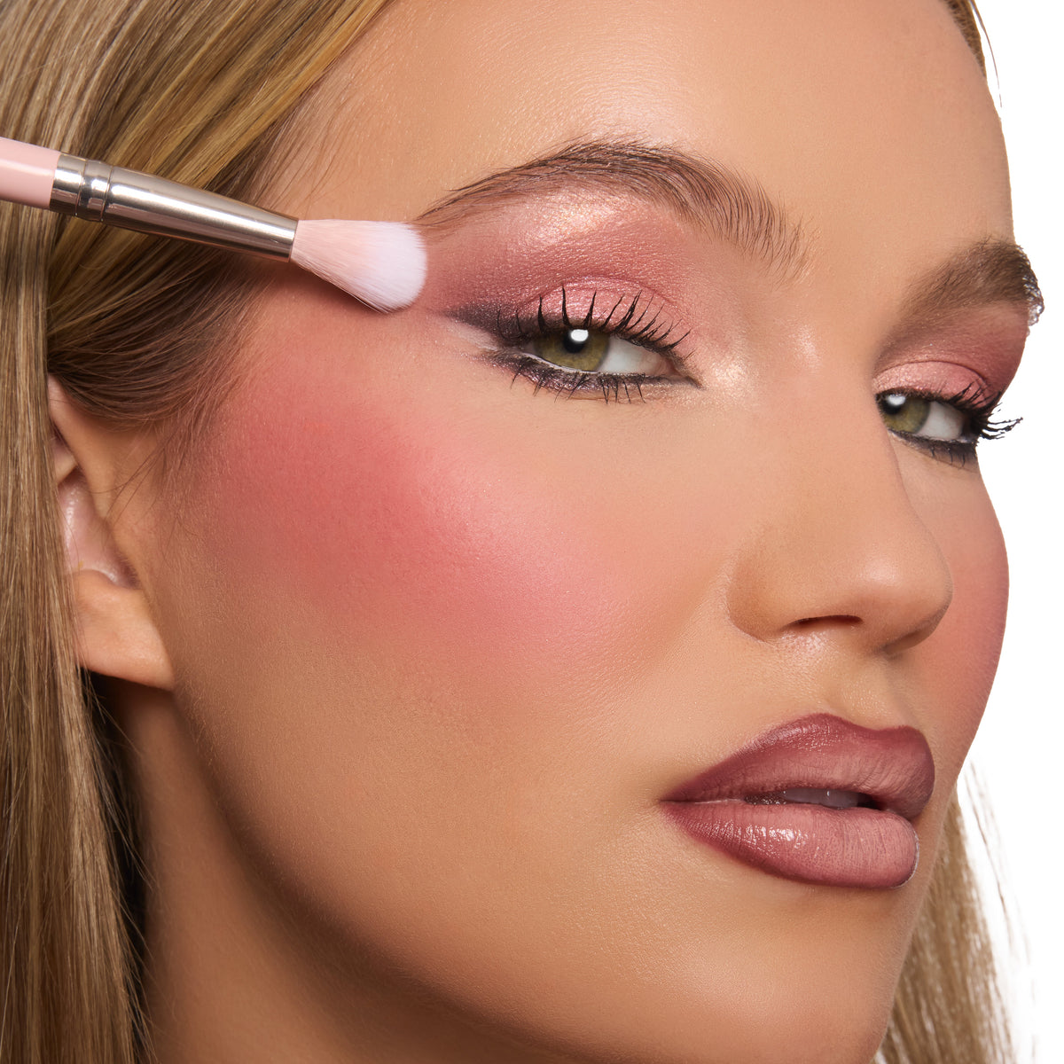 Model eye wearing rosy shimmer from the Smoke 'N Roses ColourPop Shadow Palette, makeup brush blending pink metallic shadow at the outer lid, paired with soft blush and a mauve lip, studio close-up on white background.