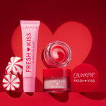 Colourpop Mistletoe Kisses lip care kit displayed upright - Fresh Kiss glossy lip balm tube, Candy Cane lip scrub jar with red lid, and swirled tinted balm pot - styled with peppermint candies against a red backdrop with a heart-shaped glow, holiday gift-ready.