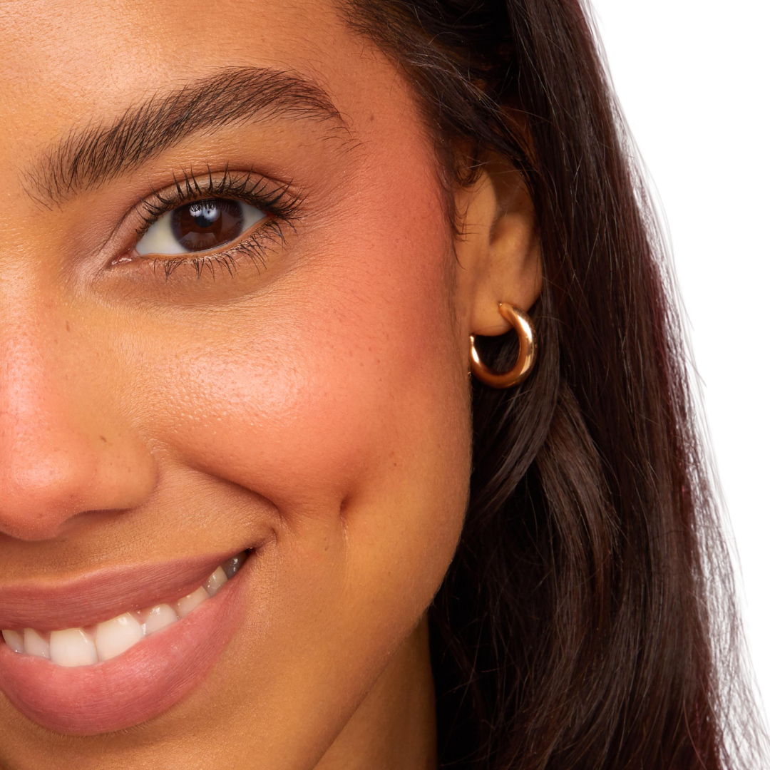 Poppi Cream Blush adding a warm peach flush to a model’s cheek, blending into natural skin for a soft, dewy finish; close-up smile with gold hoop earring on a white background, ideal to pair with ColourPop Ultra Blotted Lip.