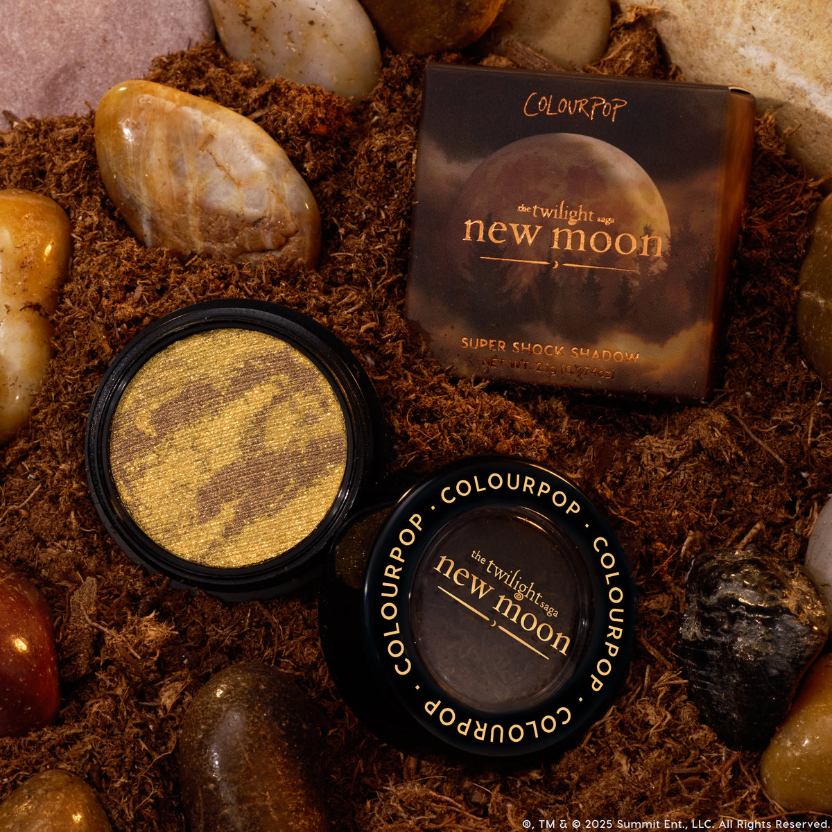 We Won’t Bite ColourPop Super Shock Shadow sits open to reveal a marbled gold and chocolate shimmer, with the closed lid and New Moon box beside it, styled top-down on forest soil with smooth river rocks from the Twilight collection.