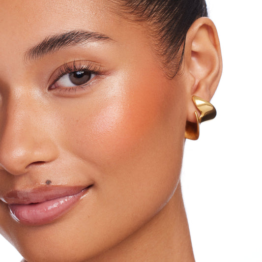 Moonstone Beach by ColourPop Bronzer Stix warming the cheekbones with a sun-kissed, dewy glow, shown in a close-up of a face with glossy nude lips and a sculptural gold hoop earring against a white background.