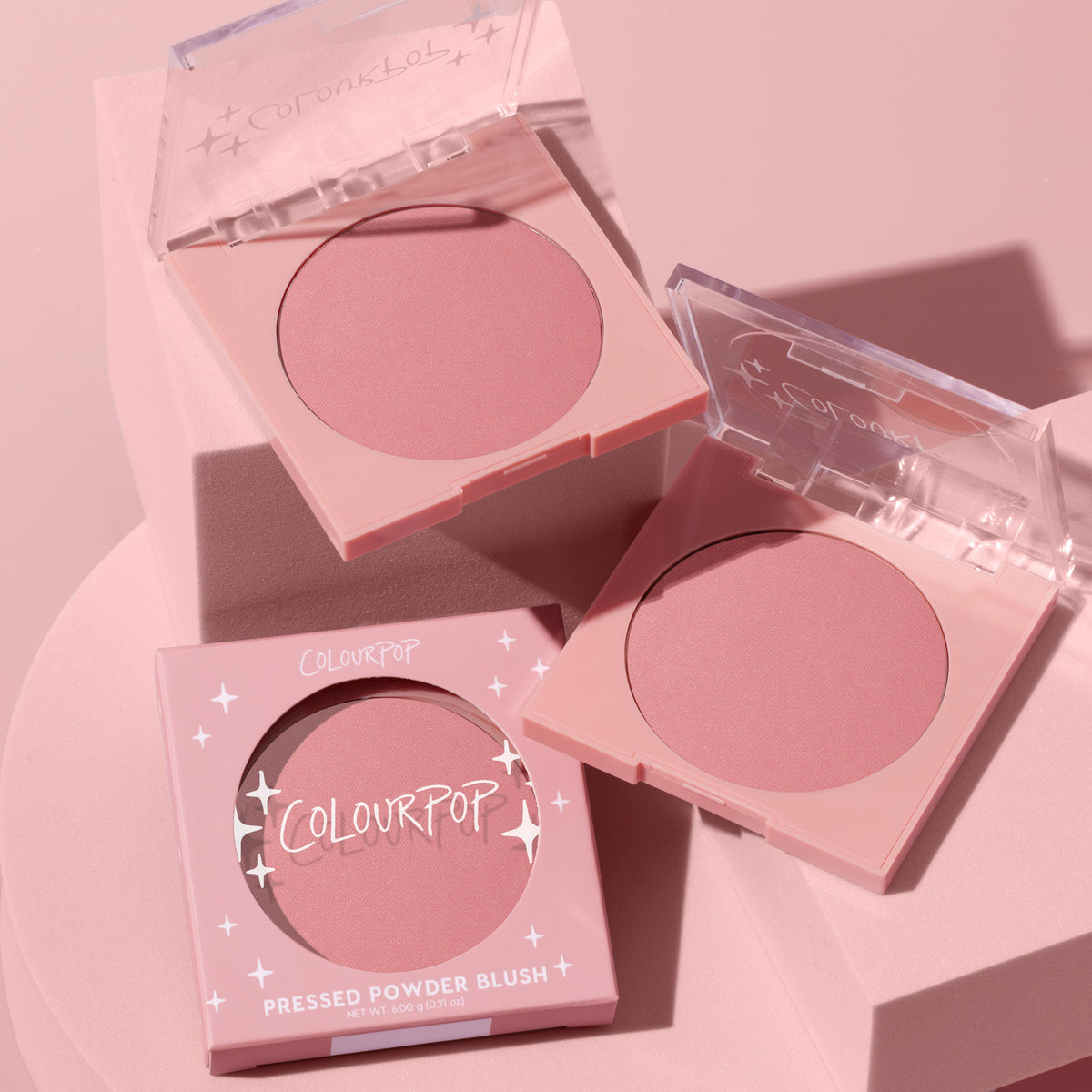 ColourPop Pressed Powder Blush in Sassy Pants, a matte dusty-rose hue, displayed in two open pastel-pink square compacts with clear lids beside the boxed unit with star accents, styled on a monochromatic blush-pink pedestal.