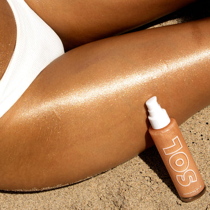 ColourPop SOL Shimmering Dry Oil in Warm Gold - pump bottle on beach sand - highlights a sun-kissed leg in a white swimsuit, with golden shimmer streaking across the skin to showcase the luminous, fast-drying body oil finish.