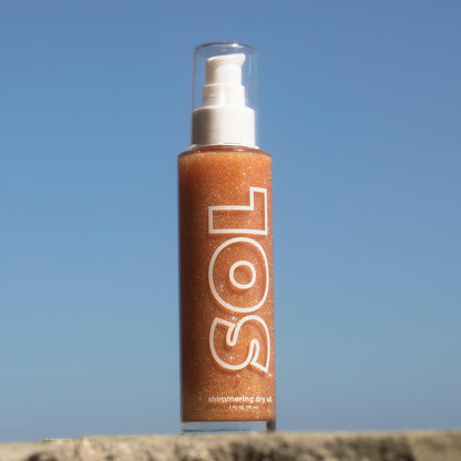 ColourPop SOL Shimmering Dry Oil in Warm Gold standing upright and sparkling in the sun, clear pump bottle with bold SOL logo, bronze-gold shimmer swirling inside, set on a stone ledge against a bright blue sky.