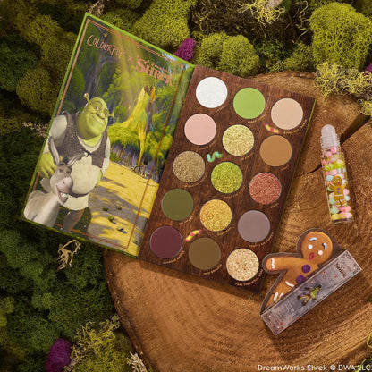 Shrek-Spress Yourself ColourPop Lip and Eye Set showcasing an open 15-pan eyeshadow palette of greens, golds, and glittery neutrals, plus a clear sprinkle lip gloss and gingerbread man accessory, styled on mossy wood for a swamp-inspired vibe.
