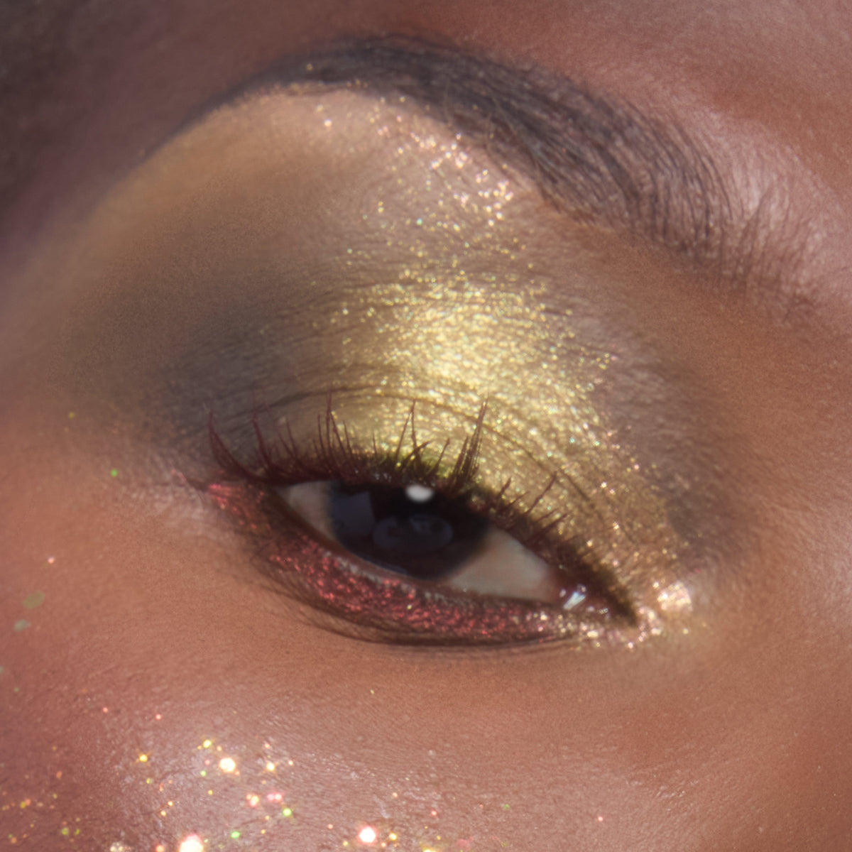 Close-up eye wears metallic lime-gold sparkle on the lid and red shimmer along the lower lashes, showcasing ColourPop Glitter Gel in Ogres are Like Onions, with chunky glitter flecks dusted across the cheekbone.