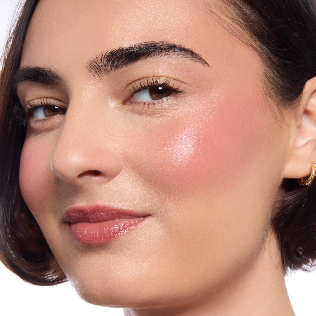 Model's cheeks and lips wear ColourPop Lip and Cheek Balm, Fairytale Things, adding a dewy rosy flush to cheeks and a soft lip tint in a studio close-up, highlighting smooth skin, natural brows, and a small gold hoop earring.