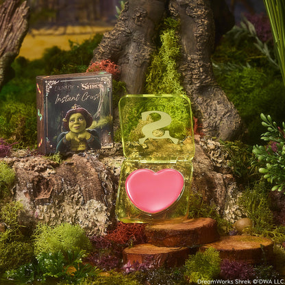 Heart-shaped pink balm sits in an open translucent lime compact with an ogre S logo, beside a box with Princess Fiona artwork, set on a mossy forest log; True Love’s Form ColourPop Lip and Cheek Balm.