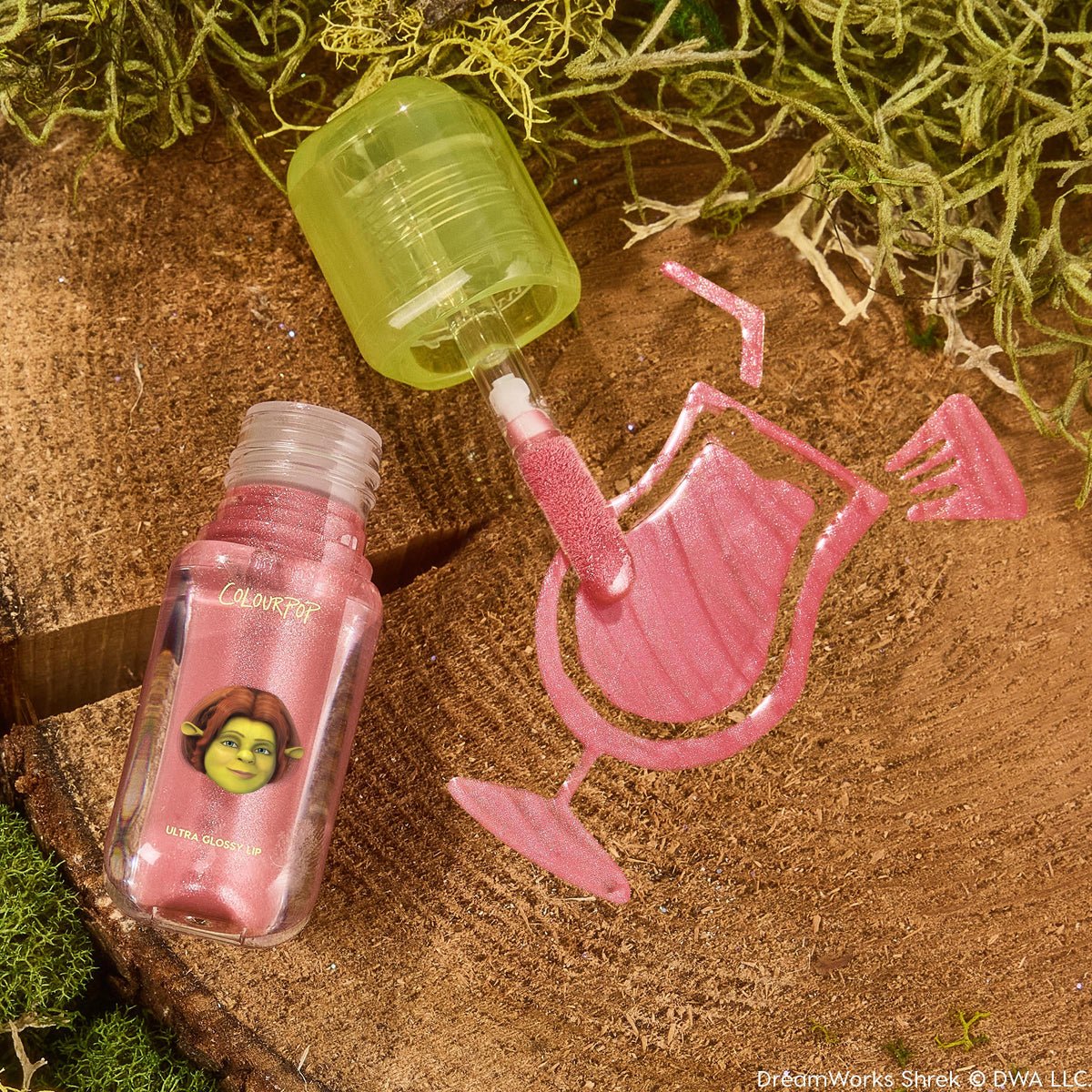 ColourPop Ultra Glossy Lip in Ogress, open with a green-capped doe-foot swiping shimmery pink gloss into a cocktail glass shape on a mossy wood stump, clear bottle showing Shrek’s Fiona artwork and a high-shine, glittery finish.