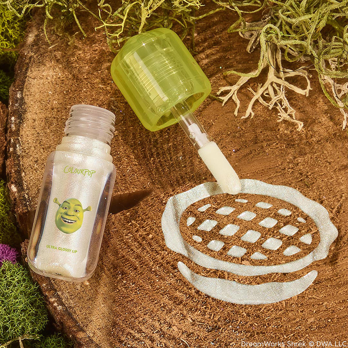 ColourPop Ultra Glossy Lip ‘Ogre’ shown open, doe‑foot applicator swiping pearlescent shimmer gloss in a swatch on a wood slice; clear tube with Shrek graphic and lime green cap, surrounded by moss for a swamp‑inspired product shot.
