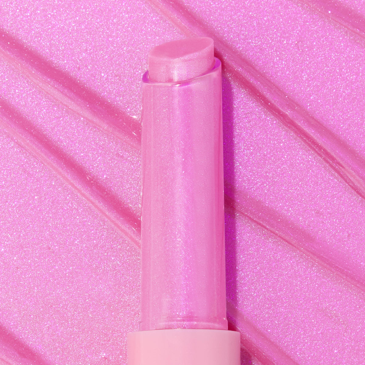 Cyberstar gloss balm’s translucent pink tube and slanted bullet standing upright, catching iridescent sparkle against streaked hot‑pink swatches; ColourPop So Juicy Plumping Gloss Balm with Peptides for glossy, plumped lips.