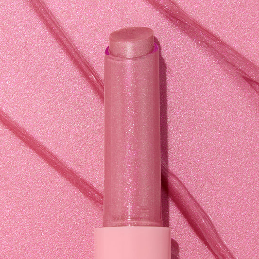 Shimmery pink Girly Pop stick of ColourPop So Juicy Plumping Gloss Balm with Peptides standing upright with the cap off, the glittery balm bullet catching light, set on a matching pink, glossy swatched background with diagonal streaks.