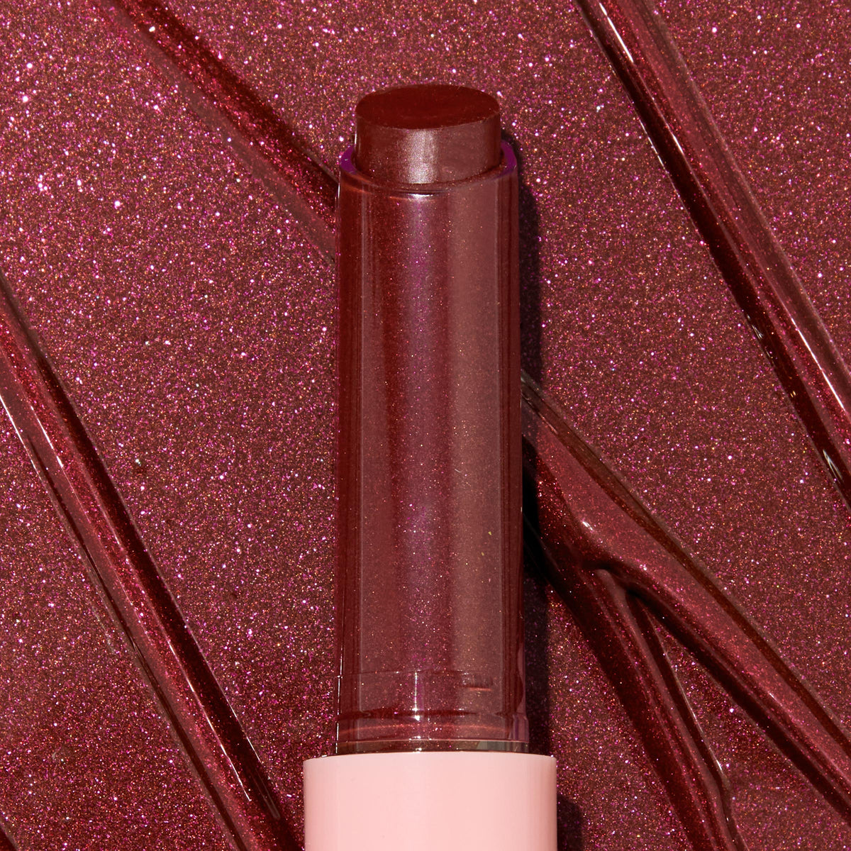 Gloss balm stick twisted up from a pink tube, L-O-V-E shade of the ColourPop So Juicy Plumping Gloss Balm with Peptides, shown close-up against glossy rose-plum shimmer swatches to highlight its dewy, plumping shine and sparkly finish.