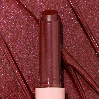 Gloss balm stick twisted up from a pink tube, L-O-V-E shade of the ColourPop So Juicy Plumping Gloss Balm with Peptides, shown close-up against glossy rose-plum shimmer swatches to highlight its dewy, plumping shine and sparkly finish.