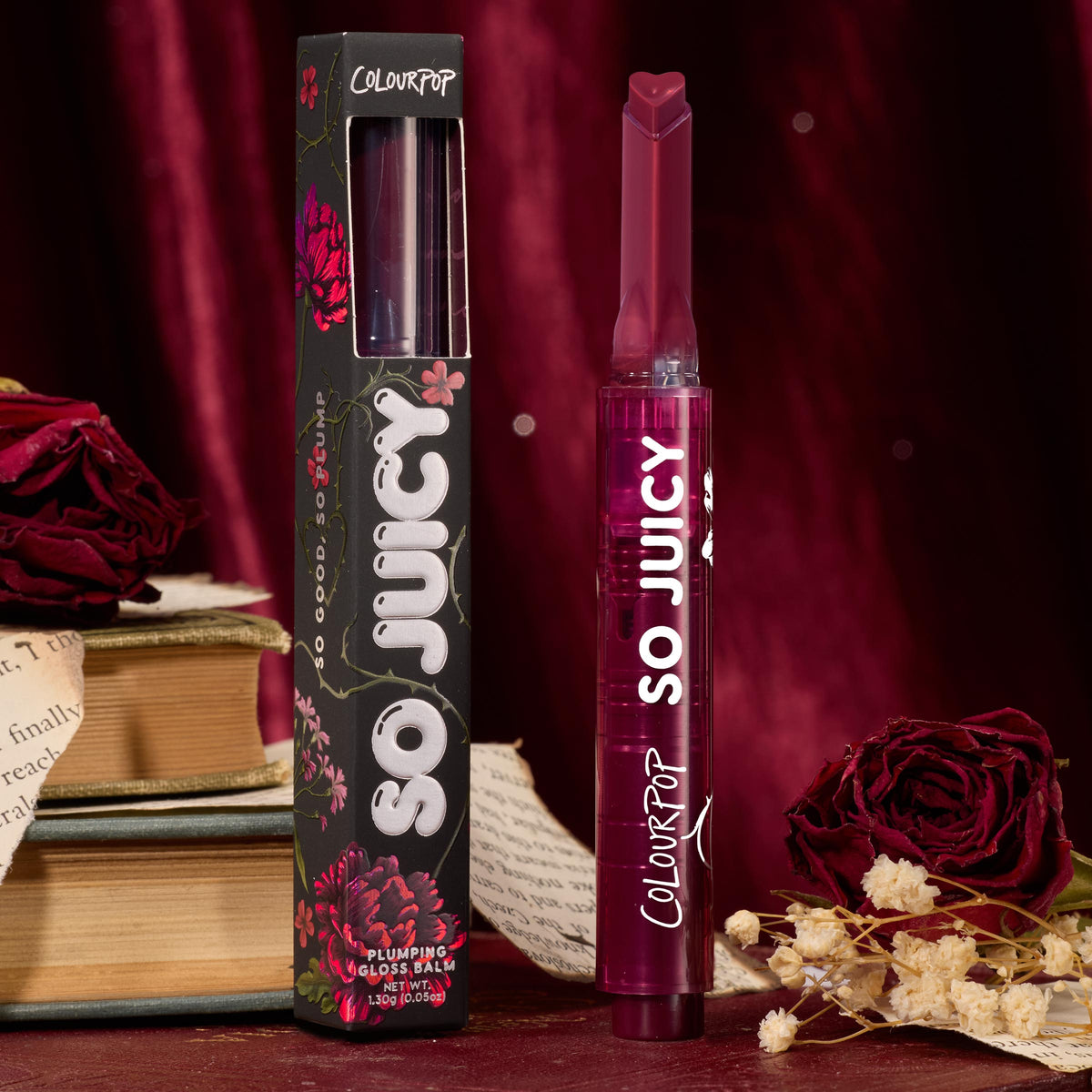 ColourPop So Juicy Plumping Gloss in Temptation stands upright with a heart-shaped tip beside its black floral box, a deep berry tube set among red velvet, roses, dried blooms, and vintage books for a romantic product shot.