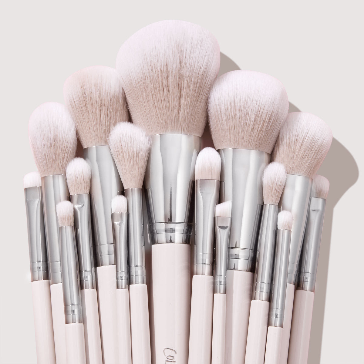 Soft pink makeup brushes with silver ferrules fan upright, showcasing powder, blush, and eyeshadow heads in a neat cluster on a neutral backdrop, part of The Ultimate Stunner ColourPop Shadow Palette & Brush Set for precise application.