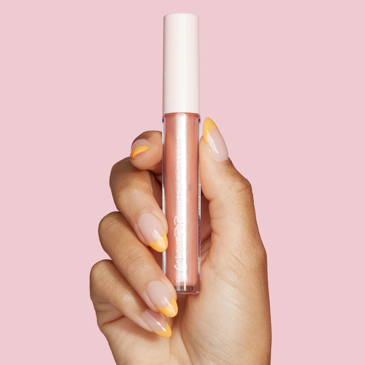 ColourPop Ultra Glossy Lip in Sugar Cookie held upright between manicured fingers, showcasing a shimmery peach‑nude gloss in a clear tube with a white star‑pattern cap and logo, against a soft pink background.