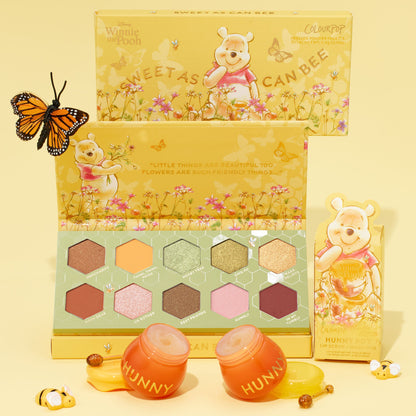 Sweet as Hunny ColourPop Eye & Lip Set shows an open Winnie the Pooh palette with 10 hexagon pans in honey-toned yellow, green, pink, and berry shades, plus two orange Hunny pot lip scrub and mask jars beside the box on a pale yellow backdrop.