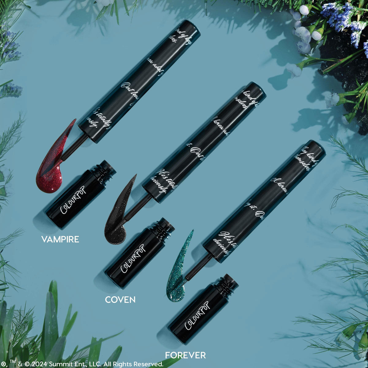 Colourpop Twilight Graphix Ink Liner I Know What You Are shown open on a blue background with foliage accents, revealing its shimmering deep red liquid eyeliner swatch next to the sleek black tube and applicator wand.