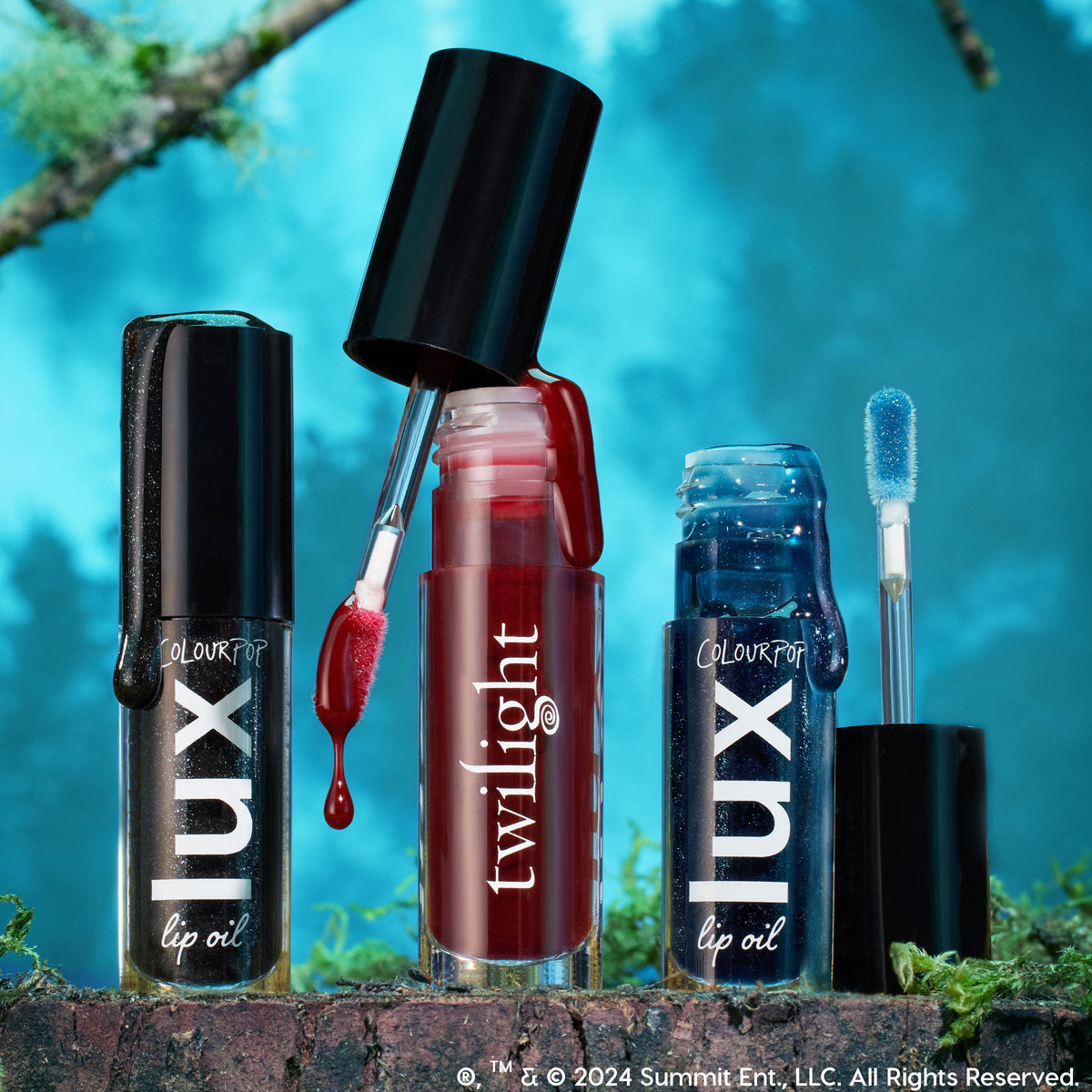 ColourPop Lux Lip Oil Vampy Lips shown with a doe-foot applicator dripping deep red gloss, flanked by black shimmer and midnight blue sparkle tubes, set on a mossy log against a teal forest backdrop.