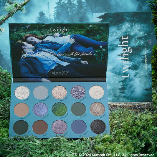 ColourPop Twilight Shadow Palette opened to reveal 15 cool‑toned matte and shimmer eyeshadows in gray, blue, green, and taupe, set on moss with the outer box; inner lid shows two characters on grass, echoing a misty forest aesthetic. ColourPop Shadow Palette.