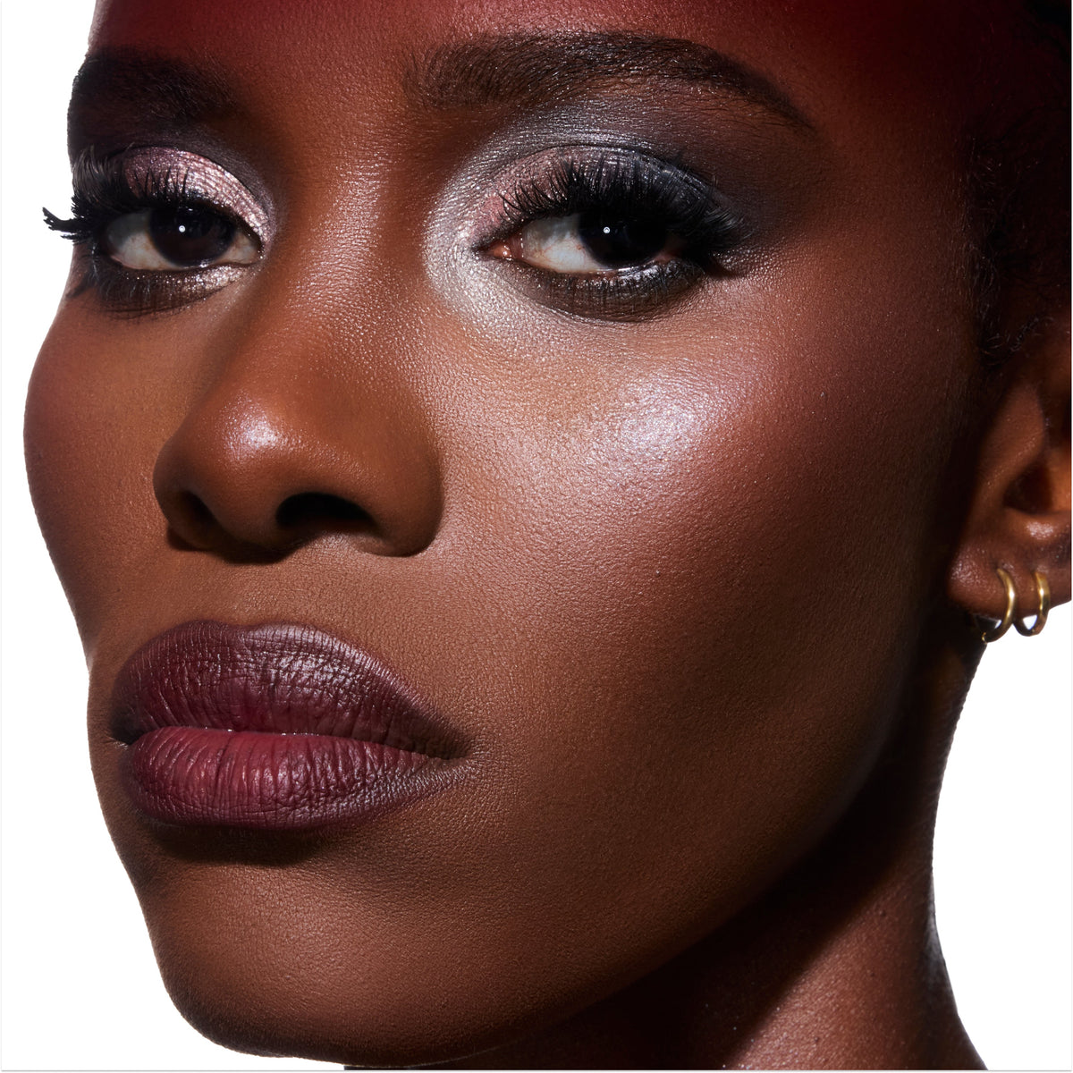 Model wearing Change Me Mega Highlighter from the Twilight collection, showing a radiant glow on deep skin with shimmering cheekbone highlight, metallic smoky eye makeup, and berry lipstick against a clean white background.