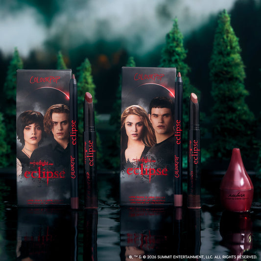 Just Bitten lip trio from the Twilight x ColourPop Eclipse collection standing upright in a dark forest setting, showing three vampy nude and berry lip pencils and sticks beside character-adorned boxes for dramatic, fandom-inspired makeup looks.