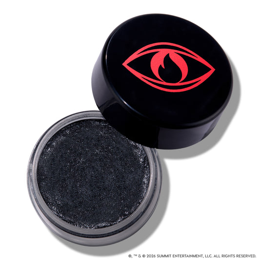 Open pot of ColourPop Newborn Army Jelly Much Shadow showing deep charcoal black, shimmer-packed cream eyeshadow, with the Twilight-inspired black lid featuring a bold red eye and flame icon resting beside it on a white background.