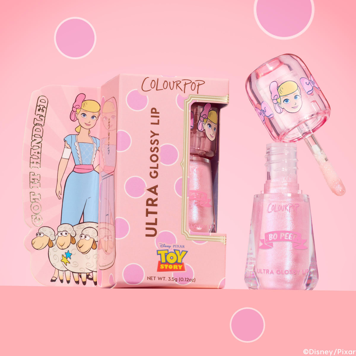 ColourPop Ultra Glossy Lip Peep This shows a shimmery light pink gloss as the wand lifts from an open clear bottle, set beside a pink polka-dot box with Bo Peep Toy Story art on a pink backdrop for a high-shine lip look.